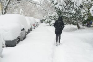 A snow-covered urban street with parked cars buried under thick snow. A person in winter clothing walks along a narrow cleared path, with snow-covered trees lining the sidewalk.