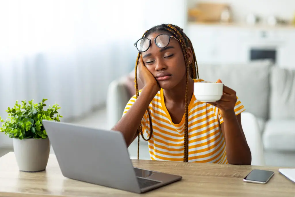Tired woman sitting at home with coffee and laptop, showing signs of burnout and fatigue.